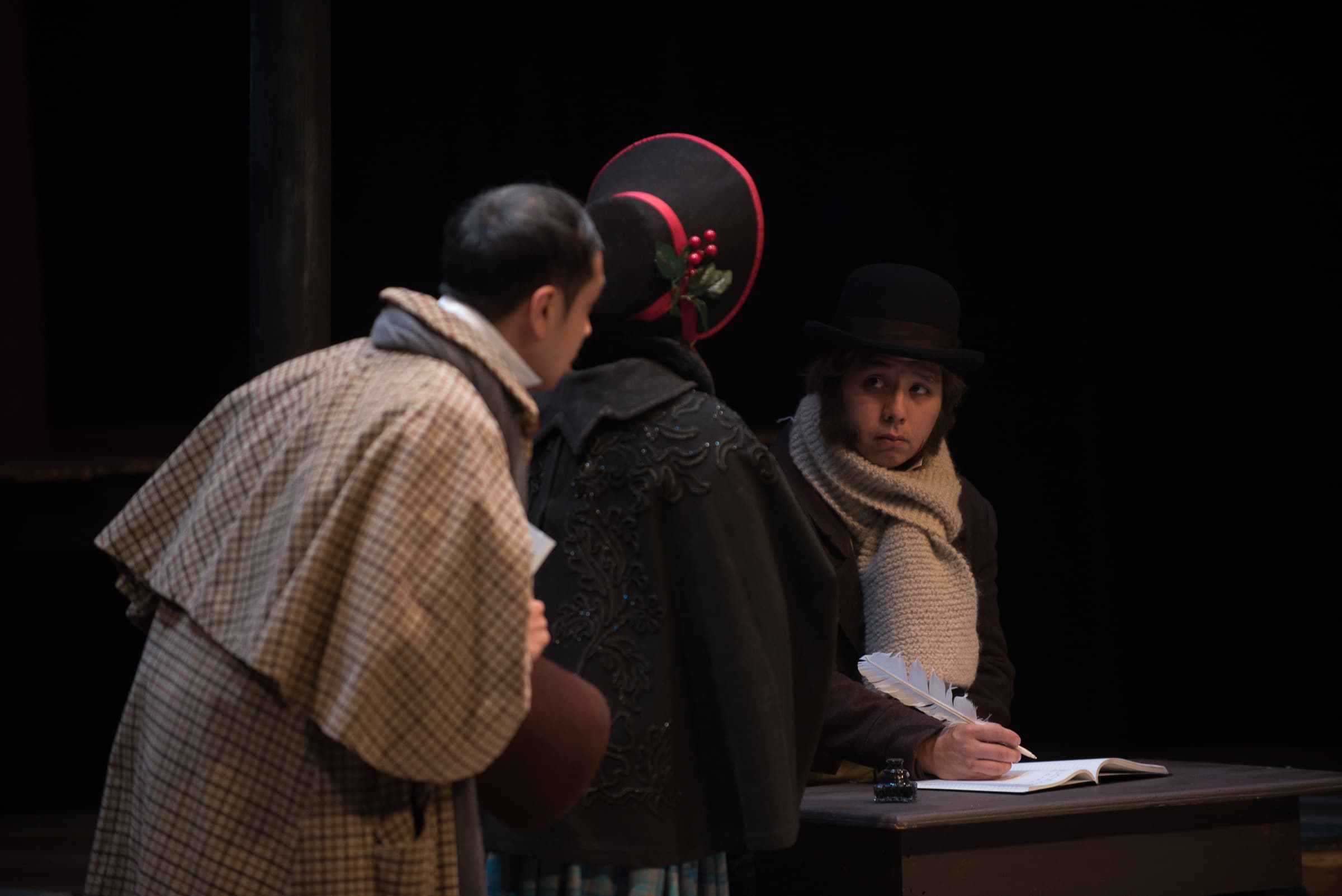 A man is on staged and dressed in turn of the century clothing and is writing with a quill while two others watch.